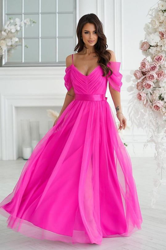 A model wearing a bright fuchsia off-the-shoulder tulle A-line gown stands holding the skirt in a white room with panelled walls a large window and a floral arrangement