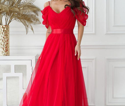 Female model wearing a red tulle off-the-shoulder A-line gown with spaghetti straps and puff sleeves standing by a white paneled wall and console table topped with a gold vase of dried foliage