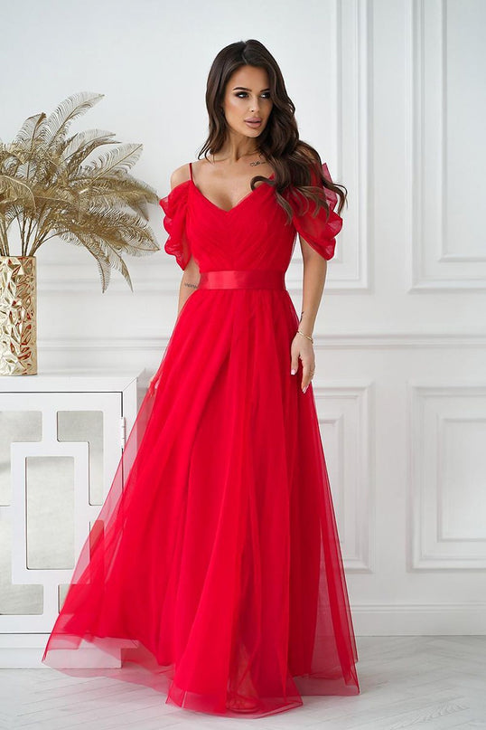 Female model wearing a red tulle off-the-shoulder A-line gown with spaghetti straps and puff sleeves standing by a white paneled wall and console table topped with a gold vase of dried foliage