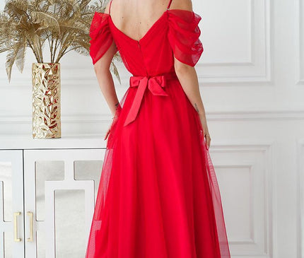 Female model wearing a red tulle off-shoulder maxi dress with spaghetti straps and waist tie facing away in a white room beside a mirrored console table topped with a gold vase of pampas grass