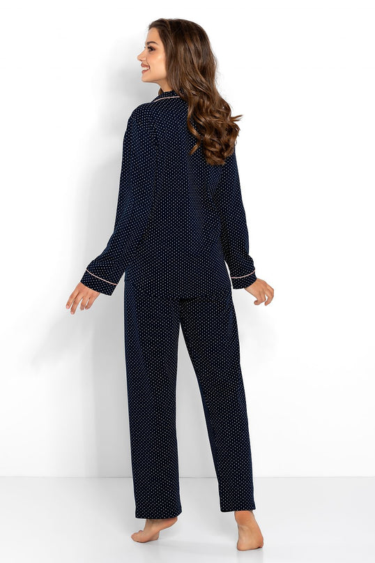 A female model stands barefoot against a white backdrop wearing a navy blue cotton polka dot pyjama set with straight-leg trousers and long sleeves while looking over her shoulder