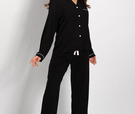 Model wears a black modal long-sleeve button-down pyjama top with white piped trim and matching wide-leg drawstring trousers standing barefoot against a plain white studio background