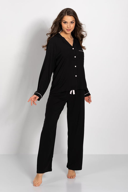 Model wears a black modal long-sleeve button-down pyjama top with white piped trim and matching wide-leg drawstring trousers standing barefoot against a plain white studio background