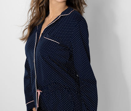 Female model in a navy blue cotton polka dot long sleeve button up pyjama top and matching shorts with pink piping stands smiling against a white studio wall