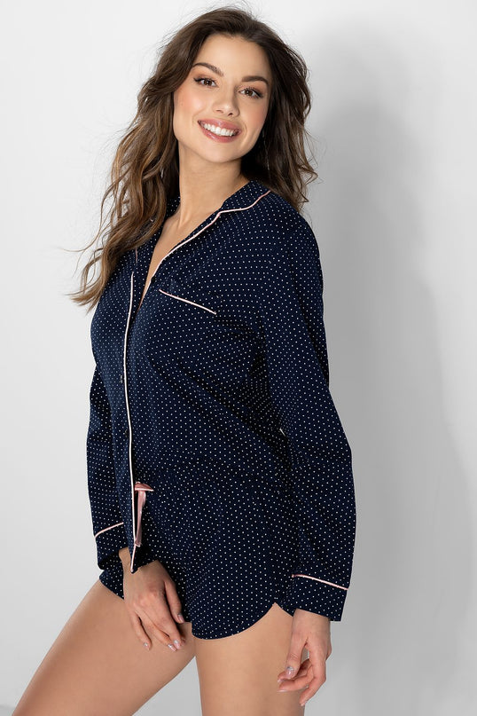 Female model in a navy blue cotton polka dot long sleeve button up pyjama top and matching shorts with pink piping stands smiling against a white studio wall