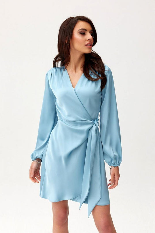 A female model stands against a plain white background wearing a light blue satin long-sleeve wrap mini dress with a V-neck and side tie looking to the side
