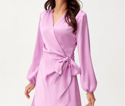 A model stands slightly turned in a white studio wearing a lilac satin wrap mini dress with long blouson sleeves a V-neckline a side bow tie and an asymmetrical hem