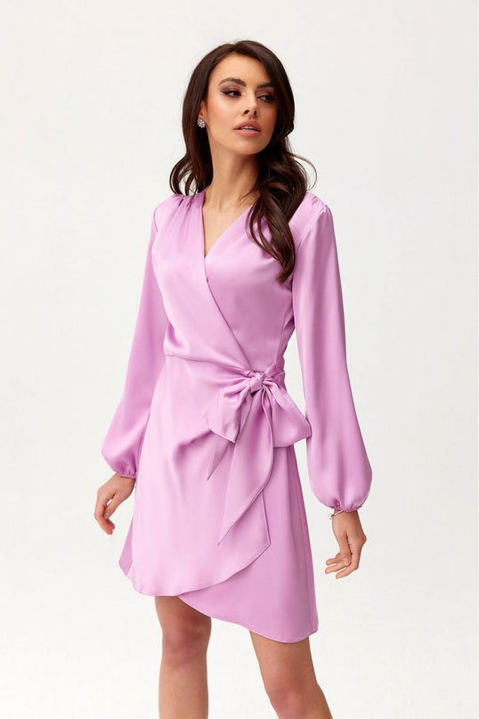 A model stands slightly turned in a white studio wearing a lilac satin wrap mini dress with long blouson sleeves a V-neckline a side bow tie and an asymmetrical hem