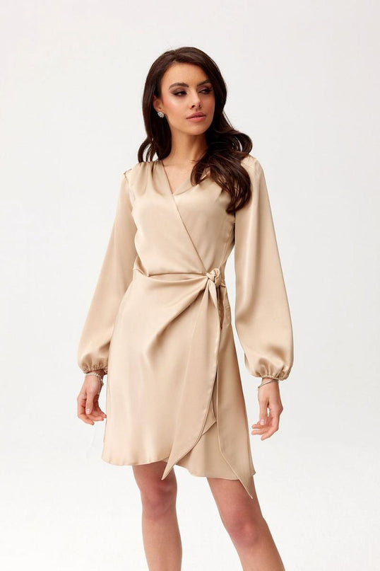 Female model wearing a beige satin wrap mini dress with long bishop sleeves stands against a plain white studio background looking to the side