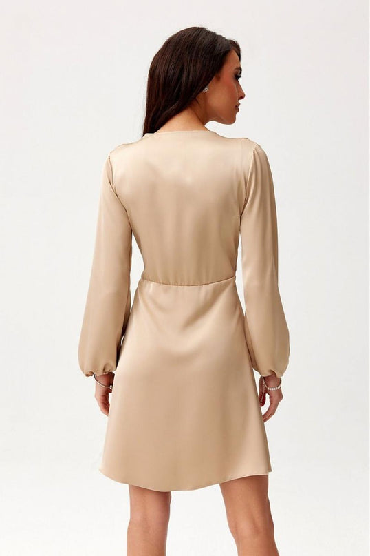 A female model stands with her back to the camera wearing a beige satin long sleeve mini dress with a flared cut against a plain white background