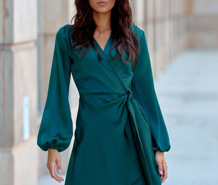 Female model wearing an emerald green satin wrap mini dress with long balloon sleeves standing in a stone columned outdoor corridor