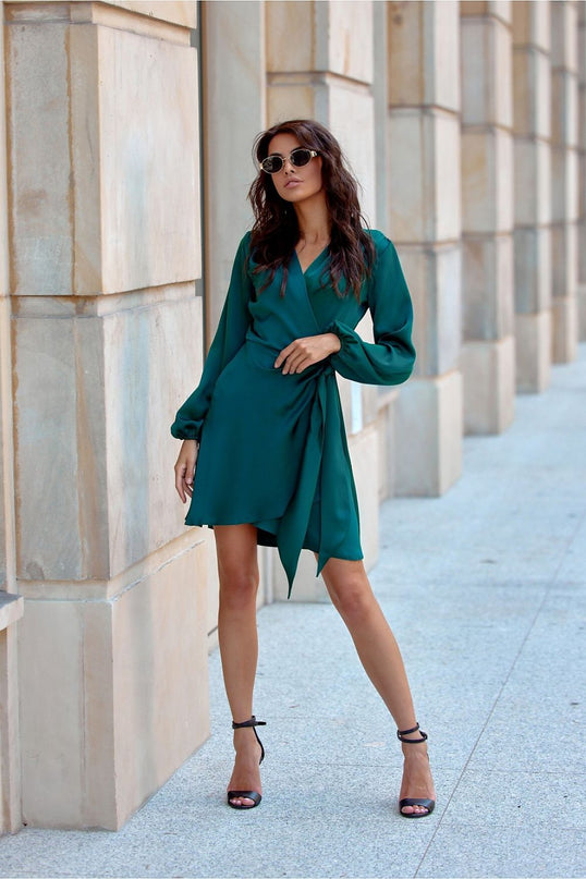 A model stands in a green satin wrap dress with long sleeves and asymmetric hem leaning against beige stone columns in a stone paved walkway