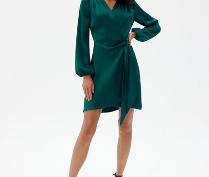 Female model stands against a white studio backdrop wearing an emerald green satin wrap mini dress with long sleeves and V-neck, posed with one hand on hip and leg slightly forward