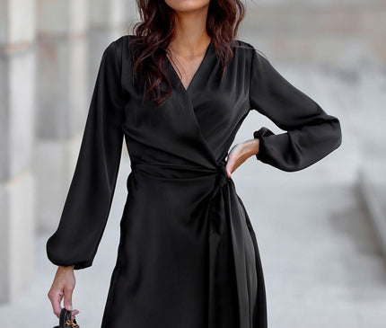 Female model wearing a black satin long-sleeve wrap dress with v-neck tie waist stands with one hand on hip and the other holding a small black bag on a grey stone pavement against a pale stone wall