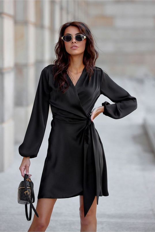 Female model wearing a black satin long-sleeve wrap dress with v-neck tie waist stands with one hand on hip and the other holding a small black bag on a grey stone pavement against a pale stone wall