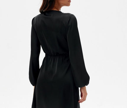 Female model in a black satin above-knee dress with long sleeves and gathered waist seen from the back with her head turned against a plain white background