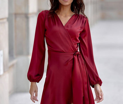 A model stands facing forward wearing a red satin wrap mini dress with long sleeves and side tie detail in a pale stone columned corridor background