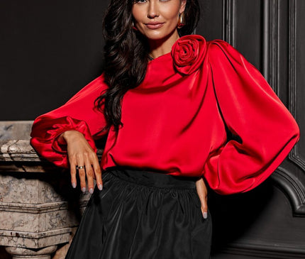 Female model in a red satin balloon-sleeved top with a rose detail and black gathered bubble skirt leaning against a marble fireplace set against a dark panelled wall