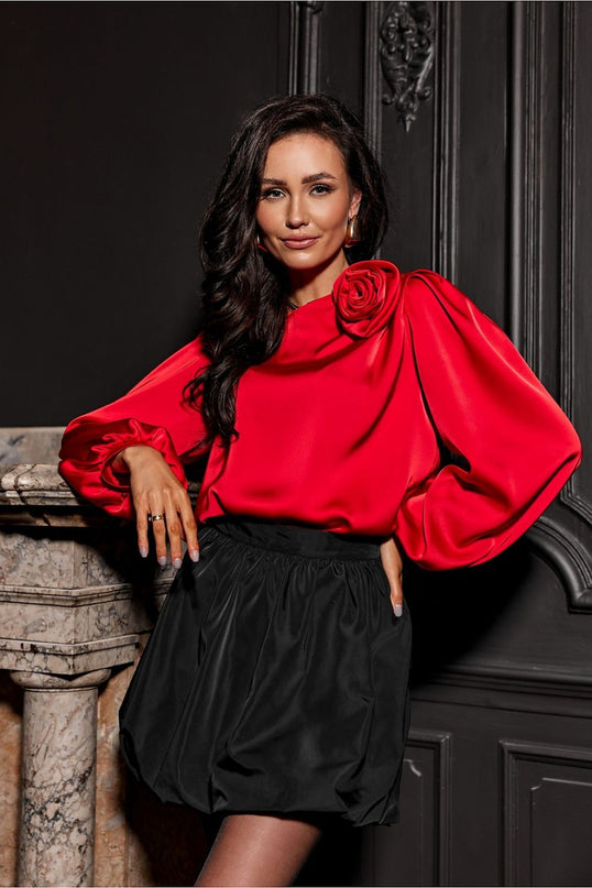 Female model in a red satin balloon-sleeved top with a rose detail and black gathered bubble skirt leaning against a marble fireplace set against a dark panelled wall
