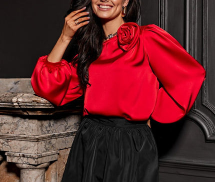 Model with hand to hair wearing a red satin puff-sleeve blouse with a rose shoulder detail and a black balloon skirt standing by a marble fireplace against a dark wood-panelled wall