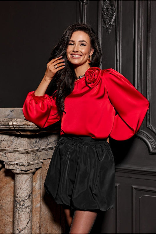 Model with hand to hair wearing a red satin puff-sleeve blouse with a rose shoulder detail and a black balloon skirt standing by a marble fireplace against a dark wood-panelled wall