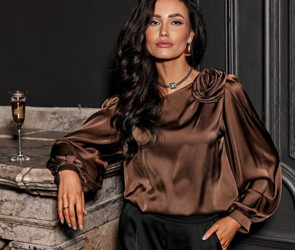 A model wearing a loose-cut brown satin blouse with rosette shoulder detail and balloon sleeves paired with black trousers leans against a marble fireplace with a champagne flute on the mantel and dark panelled walls behind