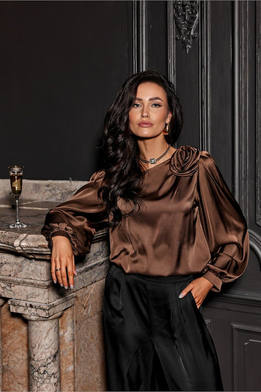 A model wearing a loose-cut brown satin blouse with rosette shoulder detail and balloon sleeves paired with black trousers leans against a marble fireplace with a champagne flute on the mantel and dark panelled walls behind