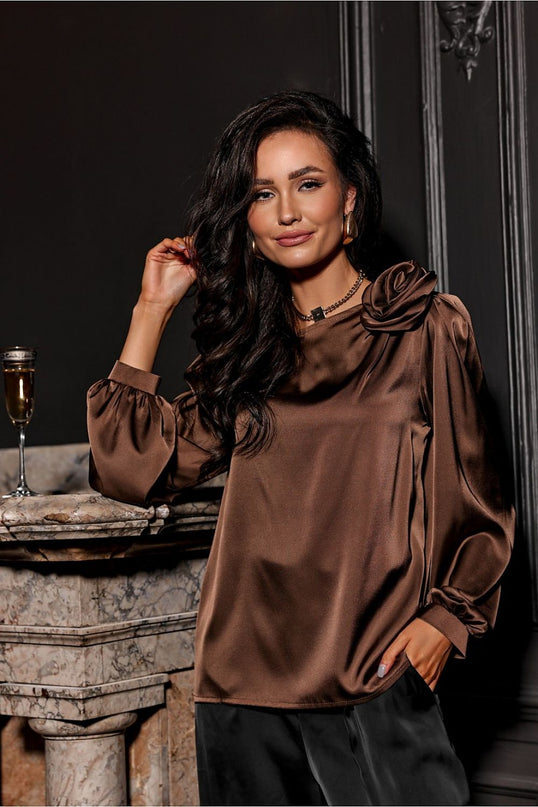Model wearing a loose brown satin blouse with gathered long sleeves and floral shoulder detail leaning against a marble mantel with a champagne glass and dark paneled wall backdrop