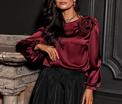 A female model wearing a burgundy satin long-sleeve blouse with rose-embellished shoulder and a black bubble skirt leans against a marble fireplace in front of a dark paneled wall