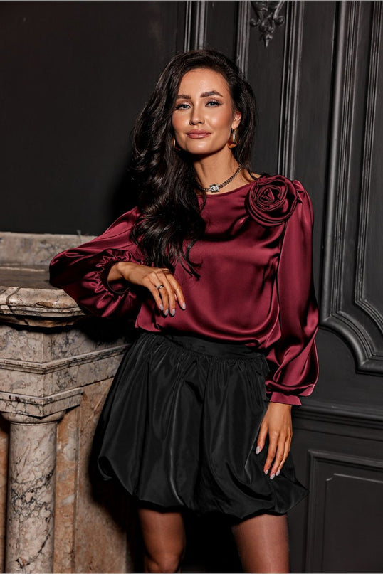 A female model wearing a burgundy satin long-sleeve blouse with rose-embellished shoulder and a black bubble skirt leans against a marble fireplace in front of a dark paneled wall