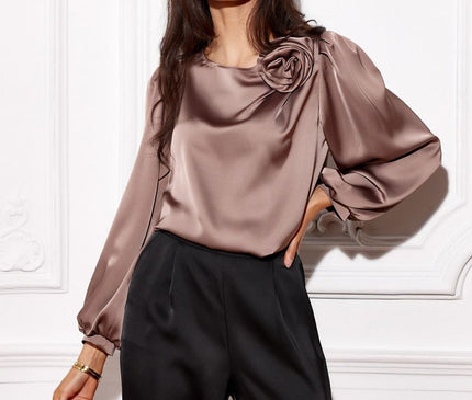 Brunette model wearing a loose-fit mauve satin blouse with balloon sleeves and rose-shaped shoulder detail posed with one hand on hip against a white moulded wall