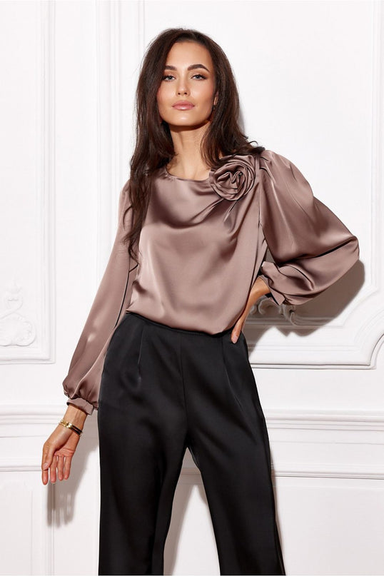 Brunette model wearing a loose-fit mauve satin blouse with balloon sleeves and rose-shaped shoulder detail posed with one hand on hip against a white moulded wall