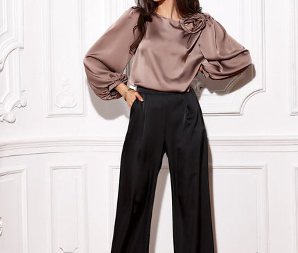 Female model with hands in pockets wears a taupe satin puff-sleeve blouse with rose detail and high-waisted black wide-leg trousers in a white panelled room with herringbone wood flooring