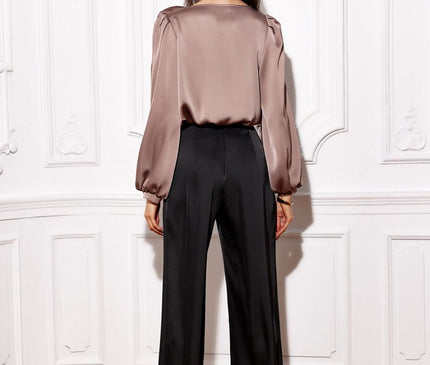 Female model stands with her back to camera and head in profile wearing a taupe satin long sleeve puffed shoulder blouse and black wide leg trousers against a white paneled wall and wooden parquet floor