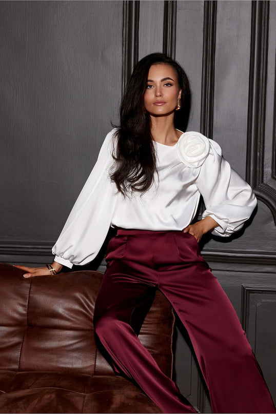Female model in a white satin balloon-sleeve blouse with an oversized rosette paired with burgundy satin wide-leg trousers leaning on a brown leather sofa against a grey paneled wall