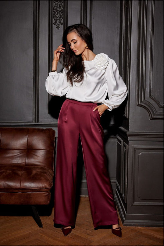 A model stands beside a brown leather sofa against a dark grey paneled wall wearing a white satin balloon-sleeve blouse with a shoulder rosette and burgundy wide-leg satin trousers