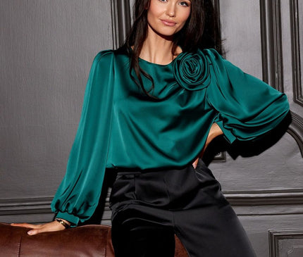 Female model wearing a teal satin blouse with rose-detail shoulder and balloon sleeves paired with black satin wide-leg trousers leans on a brown leather sofa against a grey panelled wall