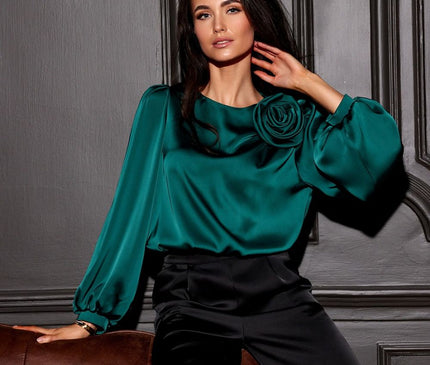 Female model wearing an emerald green satin blouse with long puff sleeves and a rose detail seated on a brown leather sofa against a grey panelled wall