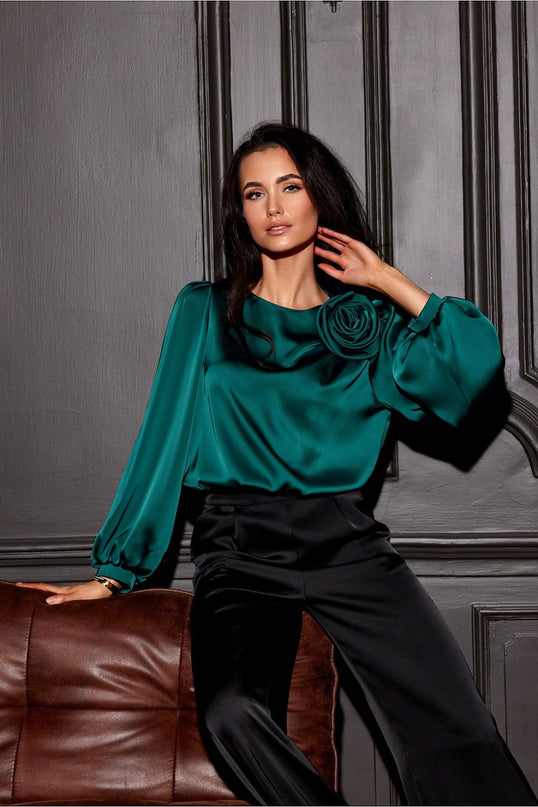 Female model wearing an emerald green satin blouse with long puff sleeves and a rose detail seated on a brown leather sofa against a grey panelled wall