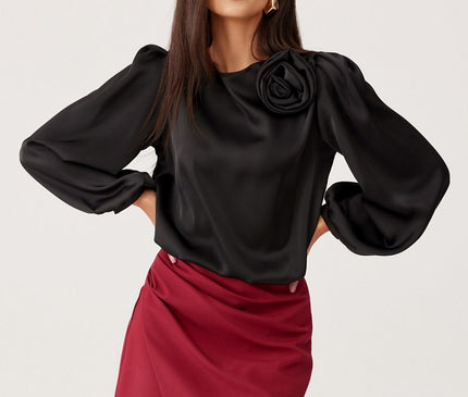 Female model stands against a white background wearing a black satin long-sleeved blouse with balloon sleeves and rose detail paired with a burgundy asymmetric wrap-front mini skirt with her hands on her hips