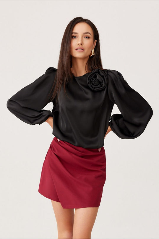Female model stands against a white background wearing a black satin long-sleeved blouse with balloon sleeves and rose detail paired with a burgundy asymmetric wrap-front mini skirt with her hands on her hips