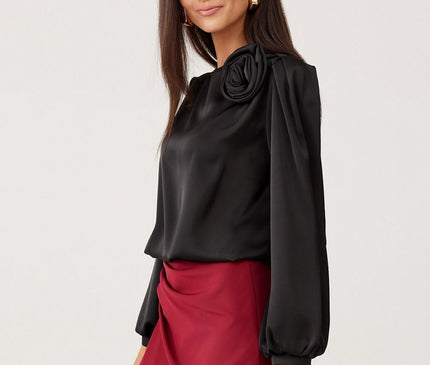Female model stands against a white backdrop wearing a black satin long-sleeve blouse with balloon sleeves and rose shoulder detail paired with a draped burgundy mini skirt