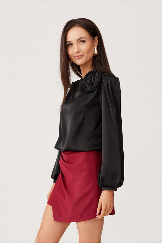 Female model stands against a white backdrop wearing a black satin long-sleeve blouse with balloon sleeves and rose shoulder detail paired with a draped burgundy mini skirt