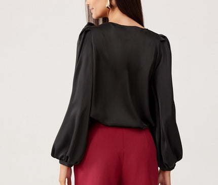 A model stands against a plain light backdrop wearing a black satin long-sleeved blouse with puff sleeves and burgundy tailored high-waisted shorts