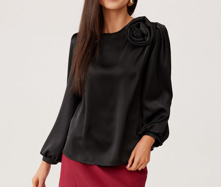 Female model stands front-facing against a plain white studio background wearing a loose-fit black satin long-sleeve blouse with a rose shoulder embellishment and a red wrap-style mini skirt smiling with one hand by her side