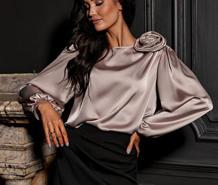 A female model in a pale blush satin loose-cut blouse with bishop sleeves and rosette shoulder detail paired with a black fitted skirt leans on a marble mantelpiece before dark ornate wall panelling