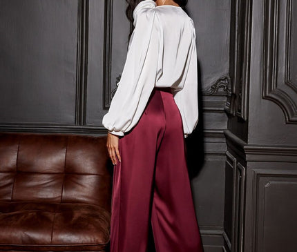 A female model wearing a white satin puff-sleeve blouse and burgundy wide-leg trousers stands looking over her shoulder against dark grey panelled walls beside a brown leather sofa on a wooden parquet floor