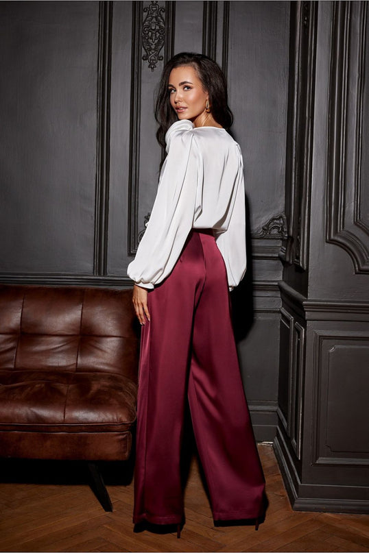 A female model wearing a white satin puff-sleeve blouse and burgundy wide-leg trousers stands looking over her shoulder against dark grey panelled walls beside a brown leather sofa on a wooden parquet floor