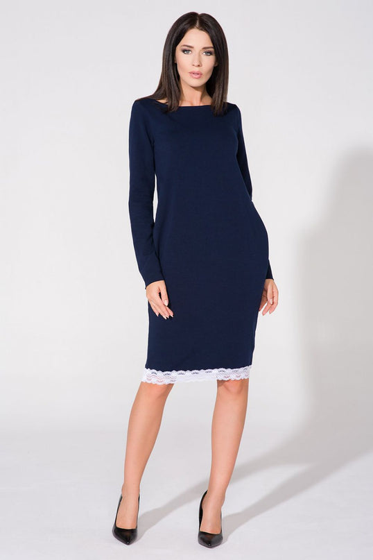 Female model wearing a navy blue knit long sleeve straight cut midi dress with white lace trim at the hem standing against a plain white studio backdrop