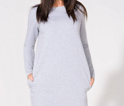 Female model wearing a light grey jersey A-line midi dress with long sleeves and black lace trim at the hem stands against a white background with her hands in pockets